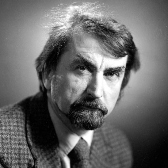 A black-and-white portrait of a man with a mustache and beard, wearing a tweed jacket and tie, looking directly at the camera with a serious expression.