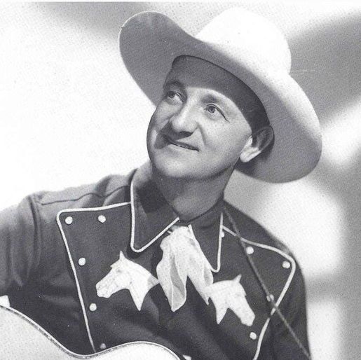 A black-and-white portrait photograph of a man smiling softly while looking upward. He is wearing a large white cowboy hat, a dark Western-style shirt with light piping along the collar and yoke, and two white embroidered horse heads on the chest. A light-colored scarf is loosely tied at the neck. He holds an acoustic guitar partially visible in the lower frame. The background is plain and light-colored with soft shadows.