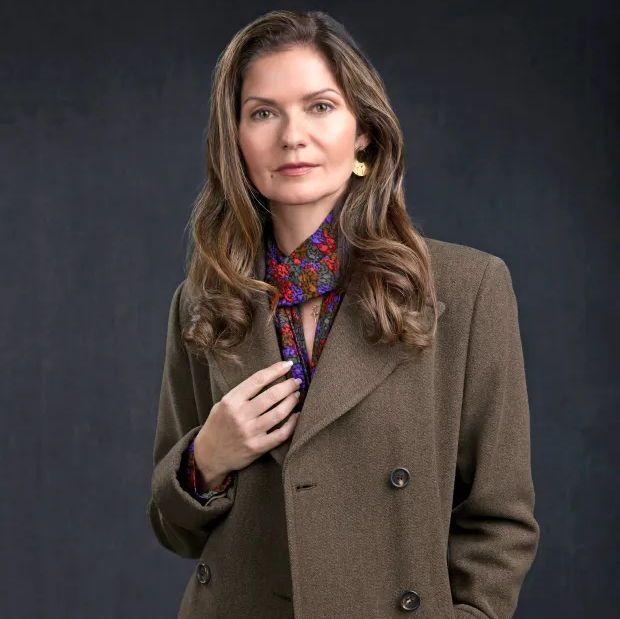 A person with long, wavy brown hair wearing a brown double-breasted coat over a colorful floral blouse, accessorized with gold earrings, stands against a dark background with one hand adjusting the collar.