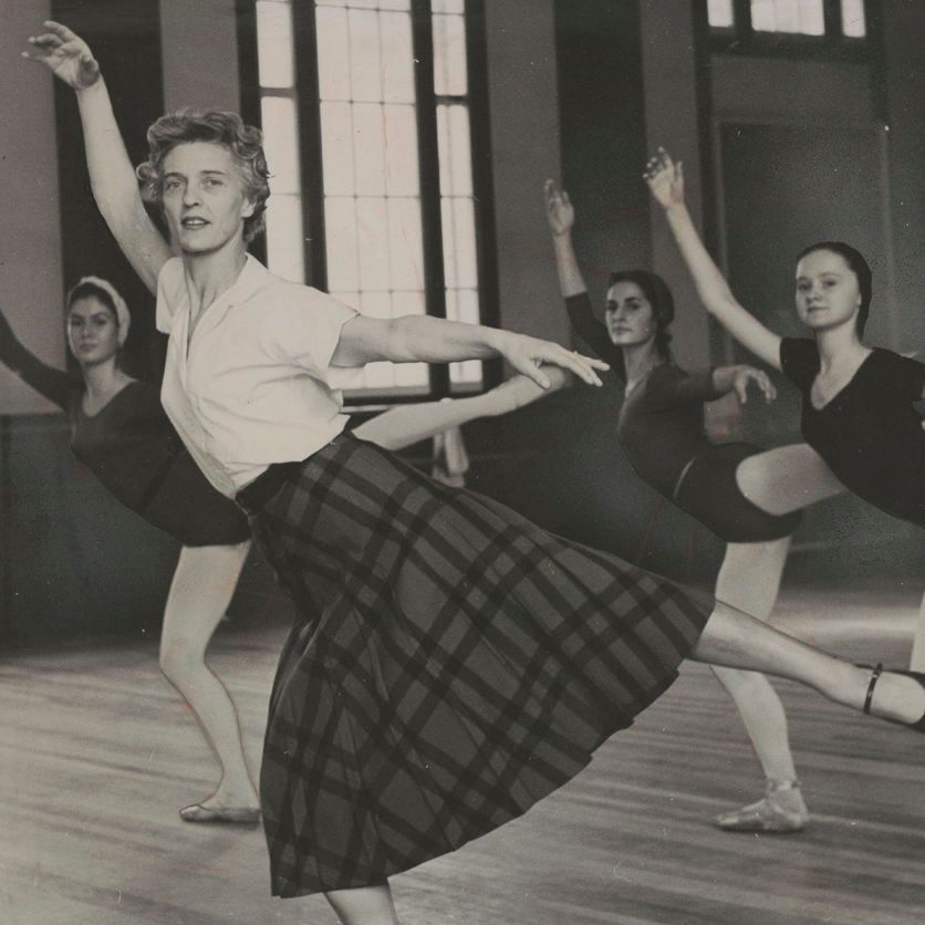 Black and white photograph of four women in a dance studio, posing with one leg extended backward and arms raised; the foreground woman wears a plaid skirt, light top, and flat shoes, while others wear leotards; arched windows in the background.