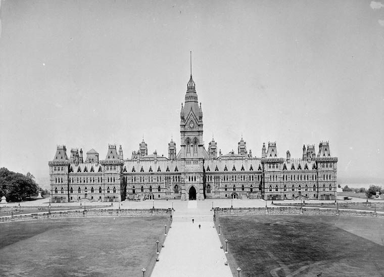 The image is a black-and-white photograph of the Parliament Buildings in Ottawa, Canada. The Gothic Revival structure features a prominent central tower with a spire, flanked by symmetrical wings with pointed arches and turrets. A wide pathway leads to the entrance, with expansive lawns on either side and a few people walking in the foreground. The photo captures a historical, grand architectural style.
