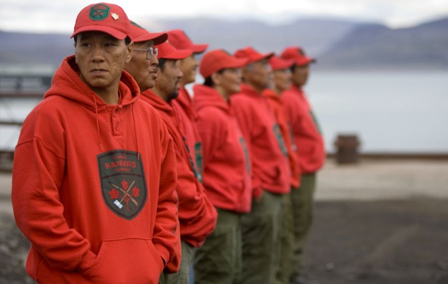 The photo shows a group of individuals standing in a line, all wearing red hoodies and matching red caps. The hoodies feature a logo with the text "CANADA RANGERS" and an emblem including crossed rifles and a maple leaf. They are dressed in green pants and appear to be in a formal or organized setting, possibly a ceremony or group gathering. The background features a scenic landscape with a body of water, mountains, and a cloudy sky, suggesting a remote or northern location. The ground they are standing on appears to be a paved or gravel surface near the water's edge.