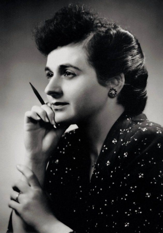 A black-and-white photo shows a person with neatly styled, voluminous hair, wearing a polka-dotted dress and earrings. They hold a pencil near their face, with one hand resting on the other arm, posing thoughtfully. The background is plain and slightly blurred.