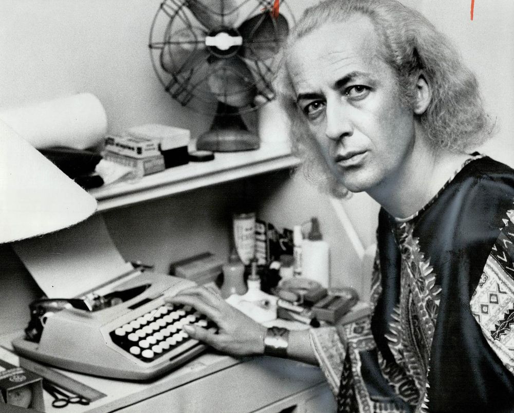 The photo, in black-and-white, shows John Herbert seated at a desk, working on a typewriter. John Herbert has long, wavy hair and is wearing a patterned garment with intricate designs, possibly a robe or tunic, which gives an artistic and unconventional vibe. His hands are on the typewriter, suggesting he is in the middle of writing, aligning with his identity as a Canadian playwright and theatre director. The desk is cluttered with various items, including a rolled-up piece of paper, a fan, a lamp, and other office supplies like bottles and containers, indicating a busy and creative workspace. A shelf above holds more papers and objects, adding to the sense of an active, working environment. The overall atmosphere reflects John Herbert's dedication to his craft, capturing a moment of focused creativity.