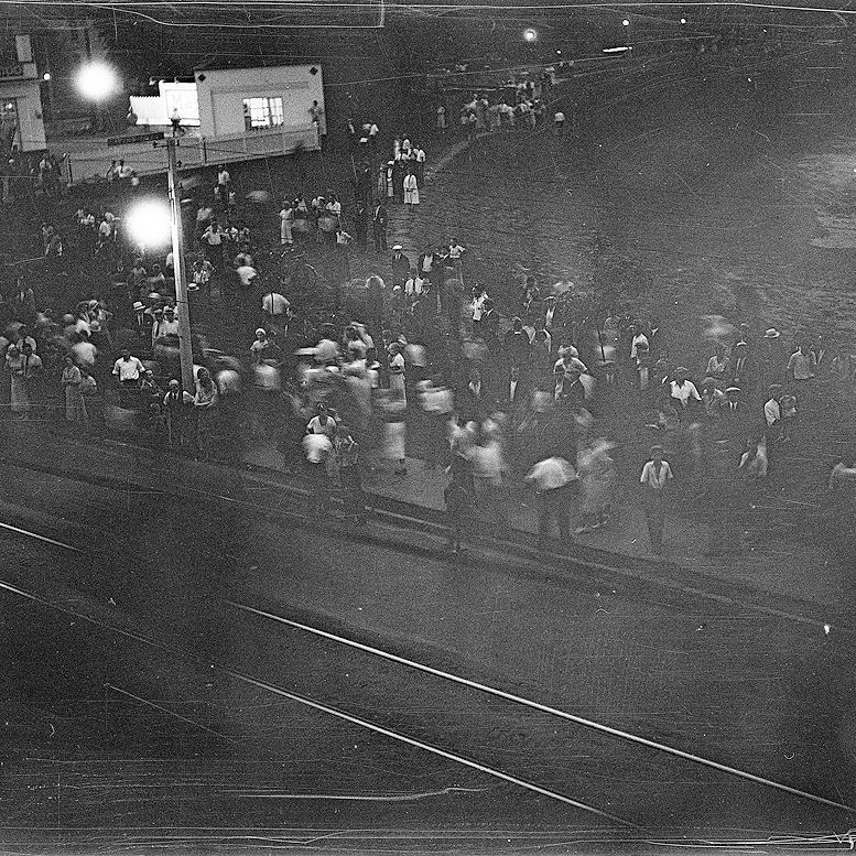 A black-and-white historical photo depicting a large crowd of people gathered on a street at night, illuminated by streetlights. The scene includes tram tracks running through the street, and the crowd appears to be moving or in motion, with some individuals blurred. Buildings and structures are visible in the background.