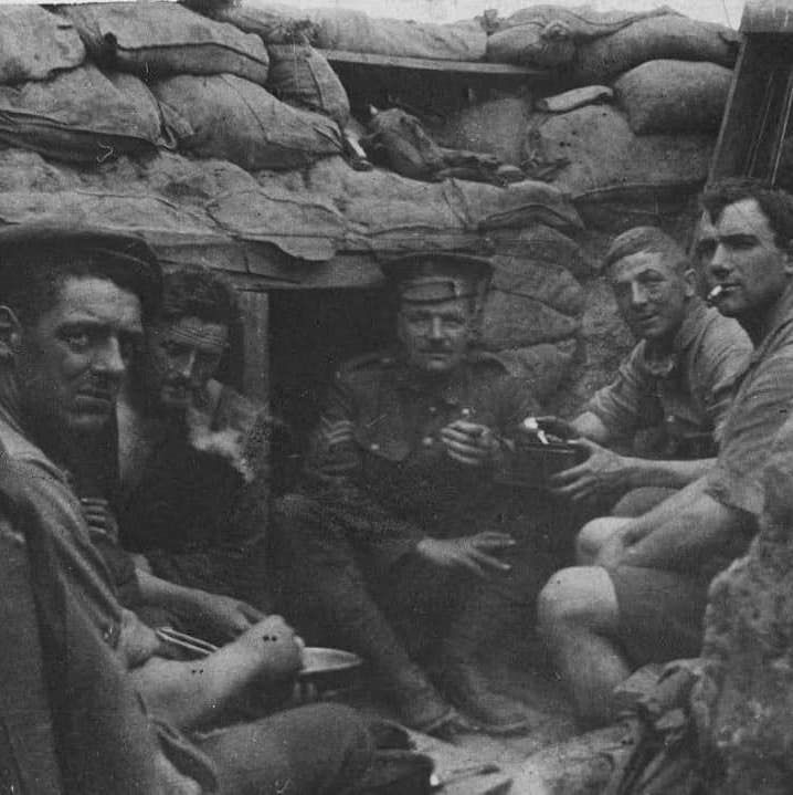 Black and white photo of five soldiers sitting inside a sandbag-walled trench, holding cups and eating, with a wooden structure above them.