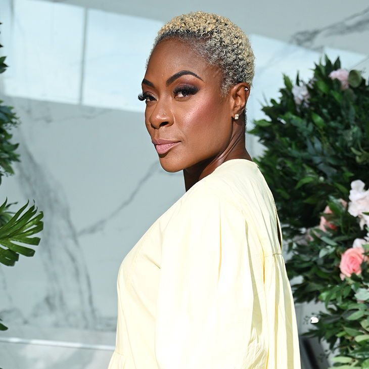 A color photo of a woman with short, platinum-blonde hair, wearing a light yellow dress, standing in front of a decorative background of lush green foliage and pink and white flowers. The setting appears to be indoors with a modern, marble-walled environment.