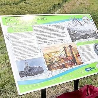 Color photograph of an elderly man with white beard, wearing a green polo shirt, khaki pants, sunglasses, and a wide-brimmed hat, standing outdoors on grass beside a large green-and-white informational sign titled "The Baptist Creek Train Disaster." He is pointing at the sign with his right hand. The sign features historical black-and-white photos of a train wreck, a bridge, a building, and a portrait, along with blocks of text. In the background, a dirt road runs alongside a field with several parked cars and a few people visible in the distance under a clear sky. A red cloth or bag lies on the ground near the sign.