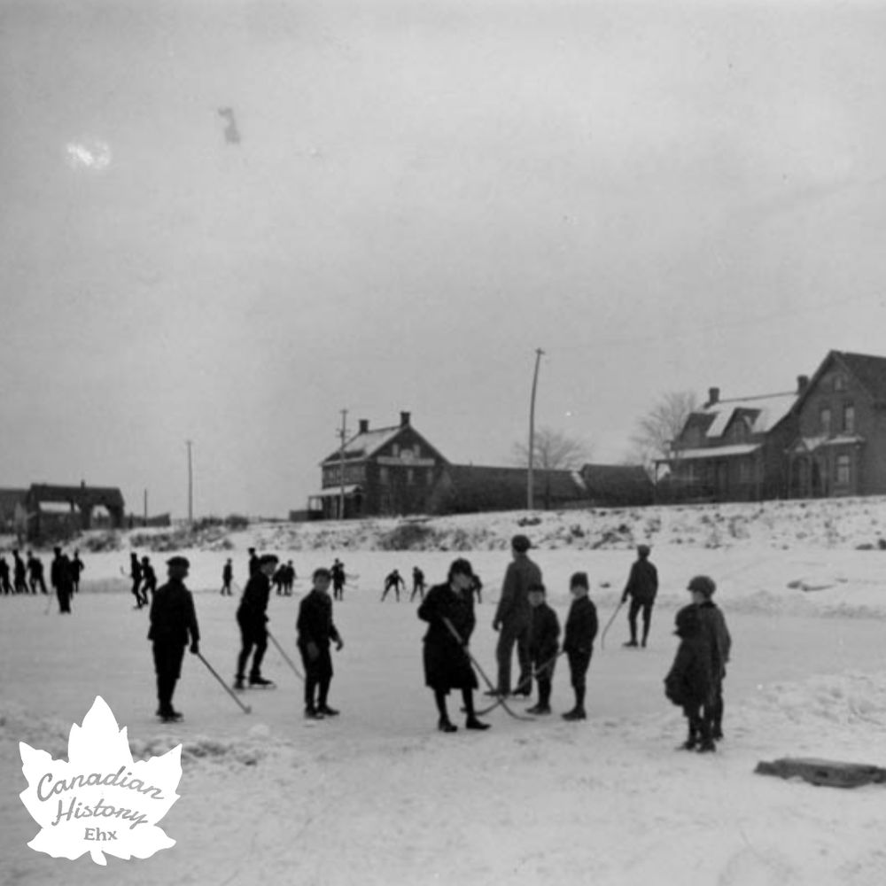This is a black-and-white photograph depicting a group of people playing hockey on an outdoor ice rink in a snowy setting. The image shows both players on the ice and spectators or players standing around the rink. The background includes houses and utility poles, indicating a residential area. The photograph has a vintage feel, suggesting it might be from the early to mid-20th century. 