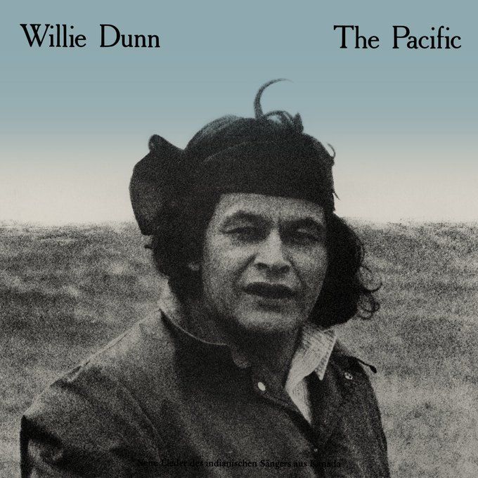 Black and white photograph of Willie Dunn, a man with medium-length wavy hair wearing a headband, looking directly at the camera with a neutral expression, dressed in a dark jacket over a collared shirt, standing in a vast open grassy field under a clear sky. The image has a blue tint gradient at the top, with text overlay "Willie Dunn" on the left and "The Pacific" on the right, and small text at the bottom reading "Some Lieder der indigenischen Sänger aus Kanada."