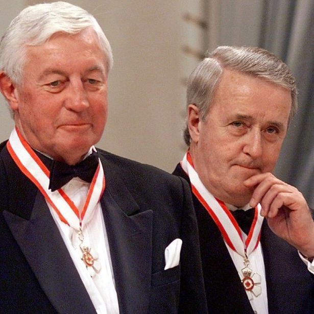 The photo shows two men in formal attire, with Jean Béliveau in the center, Brian Mulroney on the left. They are wearing black tuxedos with red and white ribbons and medals around their necks, indicating a ceremonial or award event. The background features draped curtains and a formal setting.