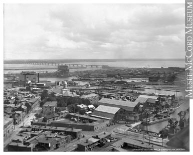 The image is a black-and-white historical photograph of an industrial waterfront cityscape, likely from the late 19th or early 20th century. It shows a dense cluster of buildings, warehouses, and docks along a river, with a long bridge spanning the water in the background. Smoke rises from chimneys, and the sky is partly cloudy, capturing an era of early urban development. The photo is credited to McCord Museum.