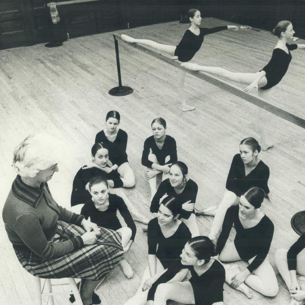 Black and white photograph of an older woman with white hair, wearing a sweater and plaid skirt, seated on a chair and gesturing while speaking to a group of young female ballet students in leotards sitting on the floor around her; two dancers perform leg extensions at the barre in the background.