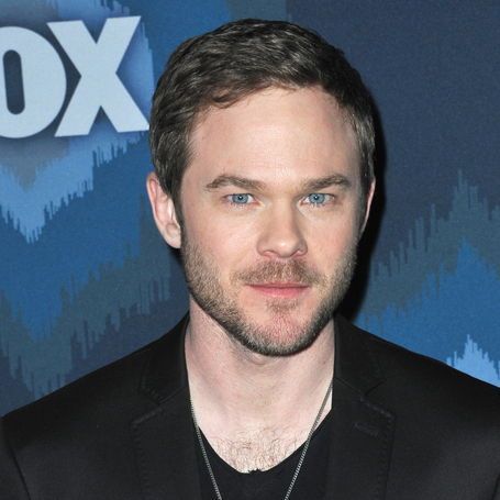 This color promotional photograph features actor Shawn Ashmore, known for playing Bobby Drake / Iceman in 20th Century Fox's X-Men film series and FBI agent Mike Weston on the Fox series *The Following*, posing against a blue backdrop with the white "FOX" logo and zigzag patterns. He has short dark brown hair, striking blue eyes, a trimmed beard, and wears a black suit jacket over a black shirt with a silver chain necklace, gazing directly at the camera with a subtle smile.