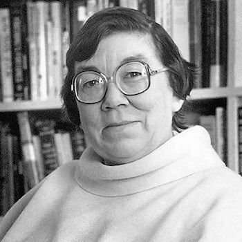 The photo is in black and white and shows a person seated in front of a bookshelf filled with books. The individual is wearing large, round glasses and a light-colored turtleneck sweater. The background suggests an indoor setting, possibly a study or library, given the extensive collection of books behind them. 
