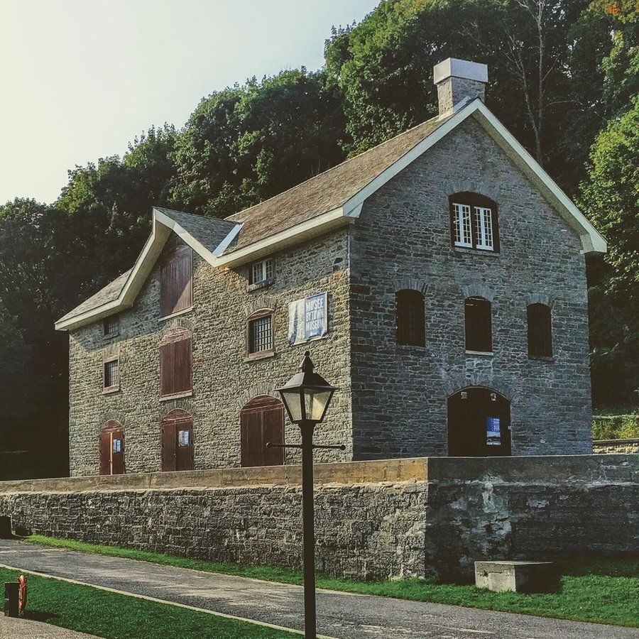 A historic stone building with a steeply pitched roof, surrounded by lush green trees, features arched windows and doors with wooden shutters. A sign reading "Museum" is visible on the facade, and a traditional street lamp stands in the foreground along a paved pathway.