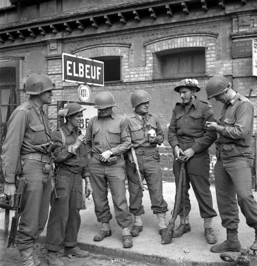This photo depicts a group of soldiers standing together in front of a building. They are dressed in military uniforms and helmets, typical of World War II era soldiers. The setting appears to be in a European town, as indicated by the sign "ELBEUF," which is a town in France. The soldiers seem to be in a moment of rest or conversation, with some holding rifles. The building behind them has a brick facade with windows, and the overall atmosphere suggests a wartime environment