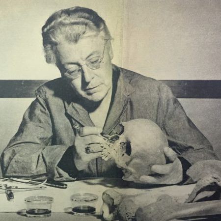 The image is a black and white photograph of a person working at a desk with scientific equipment. The person is holding a human skull and appears to be examining or working on it with tools. There are various items on the desk, including what looks like a microscope on the left, several small containers, and other scientific instruments. The setting suggests a laboratory or research environment focused on anthropology or forensic science.