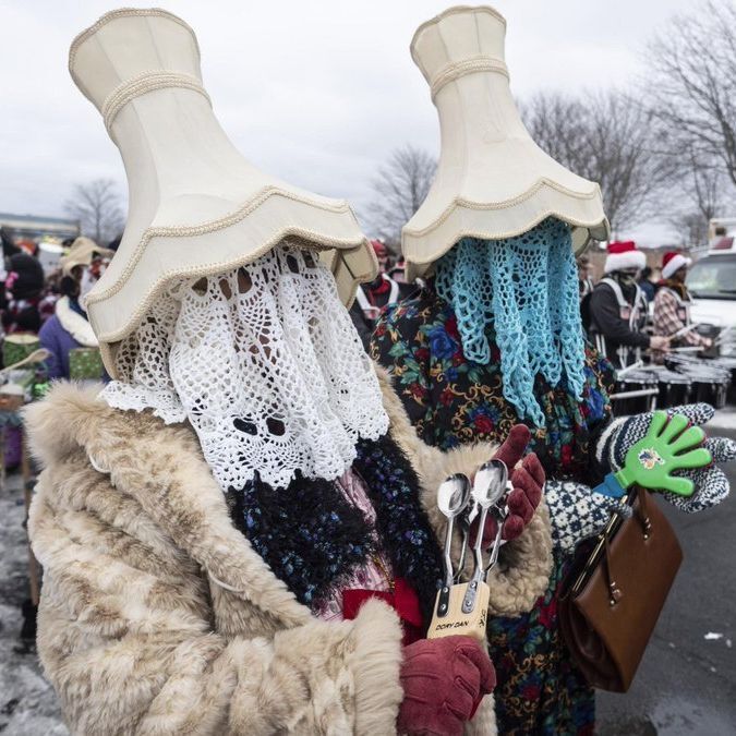 Two people stand close together outdoors in winter, both wearing large beige lampshade hats over their heads. White lace doilies hang down from the lampshades, partially covering their faces. One wears a light-colored fur coat, the other a dark floral dress with a turquoise crocheted fringe hanging from their lampshade. They hold spoons tied together with string and a brown handbag. In the background, a crowd in colorful costumes and Santa hats gathers on a snowy street with vehicles and bare trees under a gray sky.