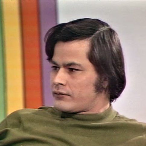 Color photograph of Willie Dunn, a man with medium-length dark hair and sideburns, gazing thoughtfully to the side with a neutral expression, wearing a green turtleneck shirt, seated against a background featuring vertical rainbow-colored stripes on the left transitioning to a white wall on the right.
