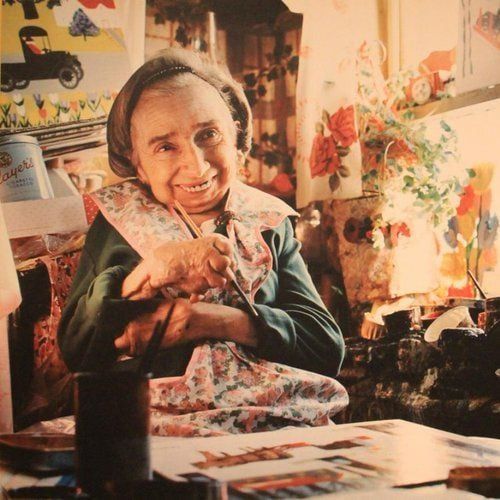 The photo depicts Maud Lewis, an elderly woman wearing a headscarf, green sweater, and floral apron, sitting at a cluttered table in a colorful room. She smiles at the camera while holding a paintbrush in her right hand, with her left arm crossed over her chest. Surrounding her are art supplies, paintings of flowers and scenes on the walls, and a basket of brushes