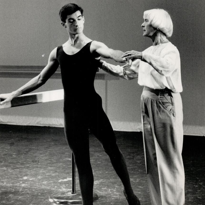 Black and white photograph of a young male dancer in a sleeveless leotard and tights practicing at a ballet barre, with an older woman in a light blouse and pants adjusting his arm position.
