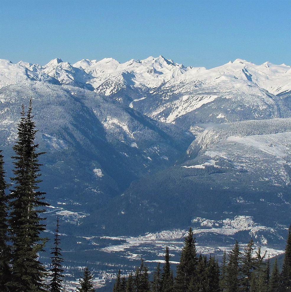 Panoramic view of snow-covered mountain peaks and ridges with a valley below containing a small town or settlement, framed by tall evergreen trees in the foreground under a clear blue sky.