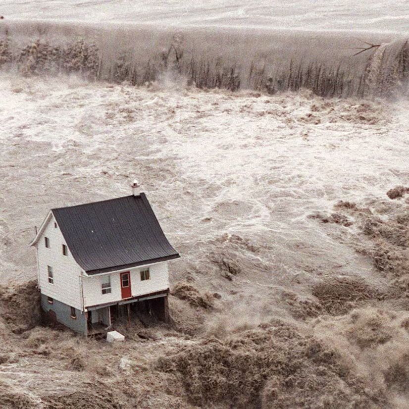 The iconic photo from the 1996 Saguenay Flood in Quebec, Canada, shows a small white house with a black roof and red door standing resiliently amid turbulent, muddy floodwaters. A massive cascading wall of brownish water looms in the background, pouring over what appears to be a breached dam or embankment, highlighting the disaster's destructive force. This structure, known as La Petite Maison Blanche, symbolizes perseverance.