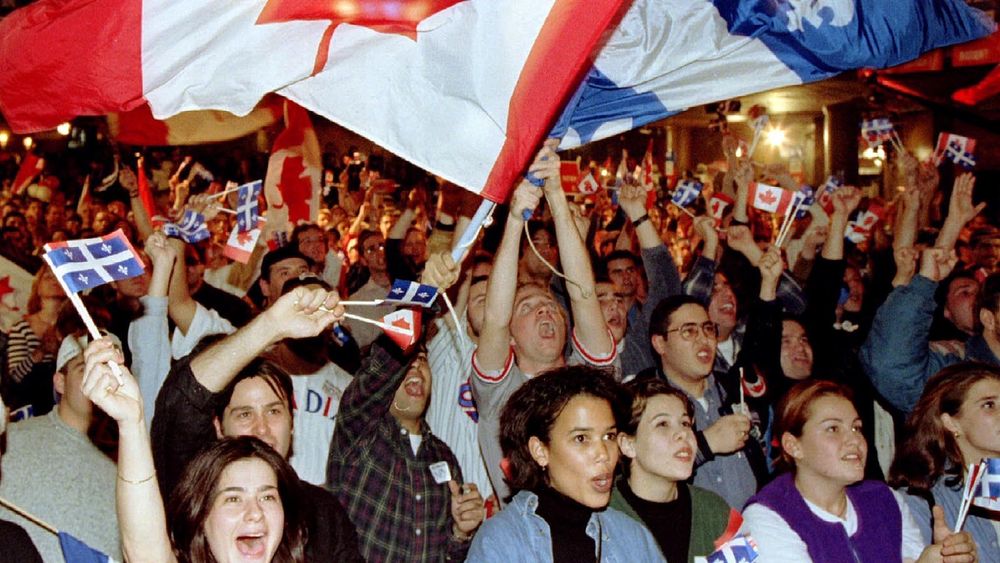 The photo shows a large crowd of people cheering and waving Canadian and Quebec flags, holding a large Canadian flag aloft. The group is in a festive indoor setting, with many raising their hands and holding small flags, creating an energetic atmosphere.