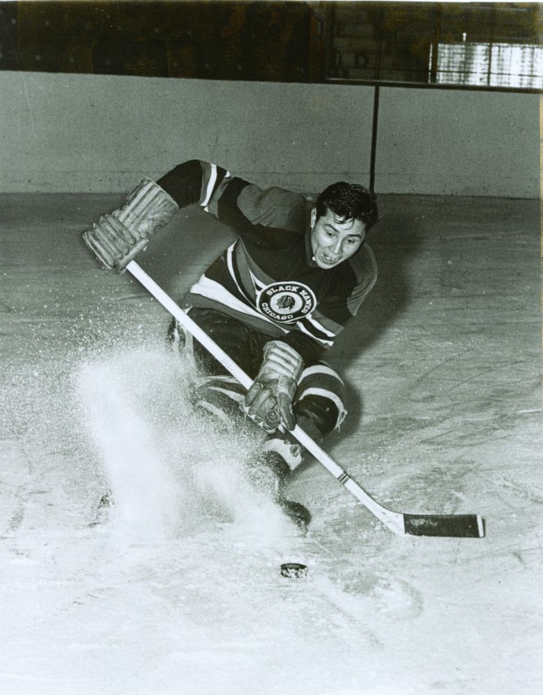 This black-and-white photograph captures a dynamic moment in an ice hockey game. The player, wearing a jersey from the "Black Hawks," is intensely focused on the puck, creating a spray of ice as he maneuvers it with his stick. The action and determination on his face, along with his posture, suggest a fast-paced and competitive game environment. The background shows the smooth surface of the ice rink, adding to the sense of motion and speed in the image.