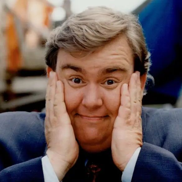 The image shows John Candy wearing a blue suit and tie, with his hands raised to his face, fingers touching his cheeks in a surprised or expressive gesture. The background appears to be an outdoor setting with blurred buildings and structures, suggesting an urban environment.