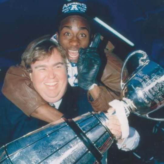 Two men excitedly pose with the Grey Cup trophy after the Toronto Argonauts' 1991 CFL championship victory. In the foreground, actor and Argonauts co-owner John Candy wears a brown leather jacket over a dark suit, white gloves, and smiles broadly while holding the large silver trophy with both hands. Behind him, wide receiver Raghib "Rocket" Ismail wears an Argonauts jersey, black gloves, and a black cap with "Argonauts" written on it, grinning widely with wide eyes and his arms wrapped around Candy's shoulders in celebration. The background is dimly lit, suggesting an indoor arena or locker room setting.