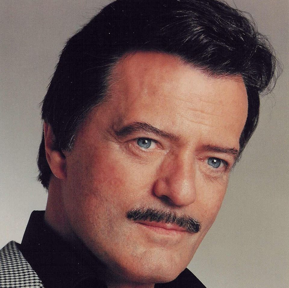A middle-aged man with dark hair streaked with gray, styled back, and a neatly trimmed mustache looks directly at the camera with a slight smile. He has light blue eyes and wears a black button-up shirt under a black-and-white houndstooth check blazer. The background is a plain light gray gradient. Studio portrait style.