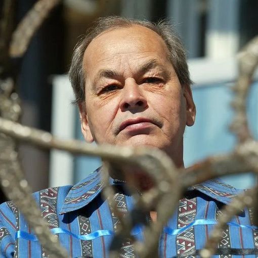 Color photograph of Willie Dunn, an older man with short graying hair, looking directly at the camera with a serious expression, wearing a blue shirt with geometric and striped patterns. He is positioned behind a foreground of twisted metal branches or fence, with a light blue building and windows in the blurred background under daylight.