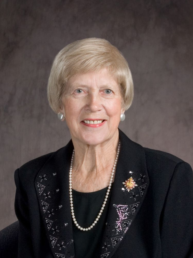 The photo depicts an individual wearing a black outfit adorned with intricate silver embroidery. The person is accessorized with a pearl necklace and matching earrings. Additionally, there is a decorative pin featuring a star and floral design on the left side of the outfit. 