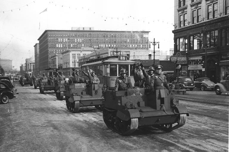 The photo is a black-and-white image of a military parade in an urban setting, likely from the mid-20th century. It shows several small tanks or armored vehicles driving down a city street, with soldiers in helmets riding in them. The street is lined with multi-story buildings, vintage cars, and streetcars, suggesting a historical context, possibly during World War II.