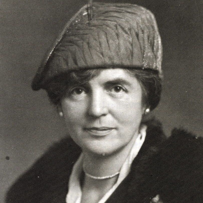 A black-and-white photograph of a woman wearing a textured beret and a dark coat with a fur collar. She has a white blouse underneath and a small floral pin on her coat. She is also wearing a necklace and pearl earrings.