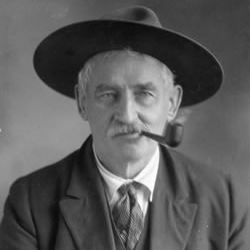 A black-and-white photograph of an older man wearing a wide-brimmed hat, a suit with a vest and tie, and smoking a pipe, with a label and code 