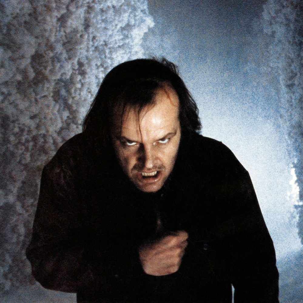 A intense close-up of Jack Nicholson as Jack Torrance from "The Shining." He has long, unkempt dark hair, a furious expression with bared teeth and glaring eyes, clutching his chest in a dark jacket. The background is a blurry, snowy tunnel or maze, evoking a frozen, menacing atmosphere.