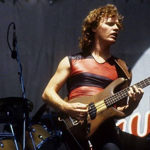 A musician with curly shoulder-length hair performs on stage, playing a brown electric bass guitar. He wears a sleeveless red and black striped shiny shirt and black pants. Behind him is a large backdrop and a drum kit is visible to his left. 