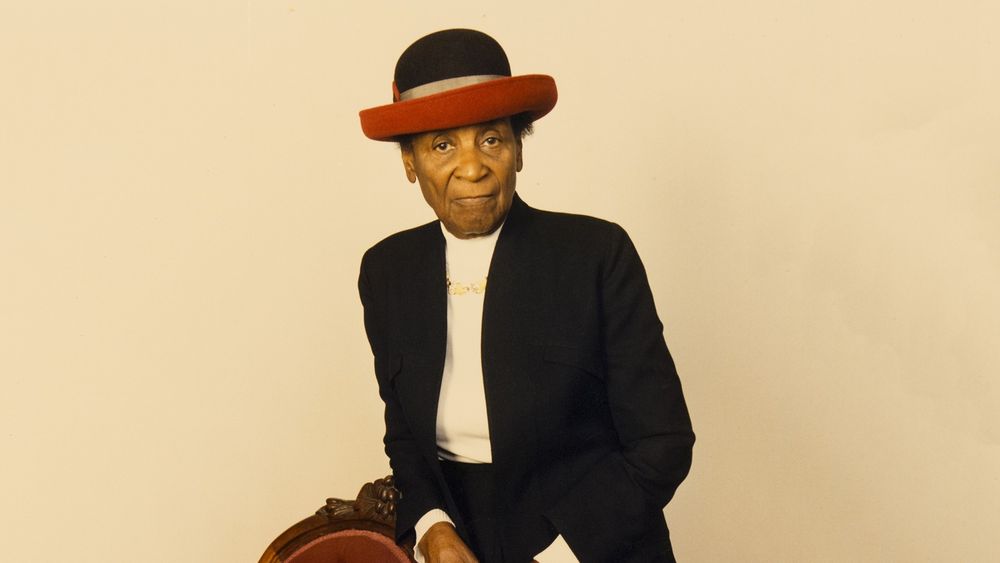 The photo features an individual dressed in a formal black suit jacket over a white shirt. They are also wearing a distinctive red hat with a black top and a gray band. The person has one hand resting on a piece of furniture, possibly a chair, which has an ornate design with what looks like carved details. The background is a plain, light-colored wall, which keeps the focus on the individual's attire and posture. The overall setting suggests a formal or possibly a staged portrait environment.