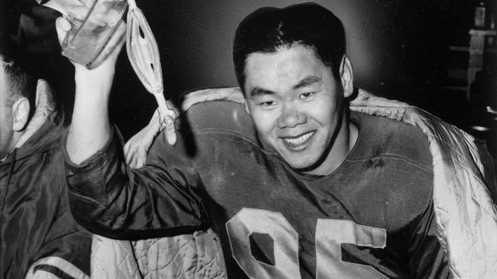 This black-and-white photo shows Norman Kwong, a football player, wearing a jersey with the number 95. He is holding a football helmet in one hand and has a quilted jacket draped over his shoulders, smiling broadly in what appears to be a celebratory moment, possibly after a game. The background is dark, with other figures partially visible.