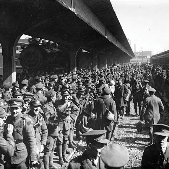 Black-and-white photograph showing a dense crowd of men, mostly wearing military uniforms with peaked caps, some carrying rifles or bags, standing and walking along a gravel-covered railway platform between two parallel trains under a long overhead canopy supported by arches. A steam locomotive is positioned at the front of the left train, with passenger cars extending into the distance where more people and industrial structures are visible.