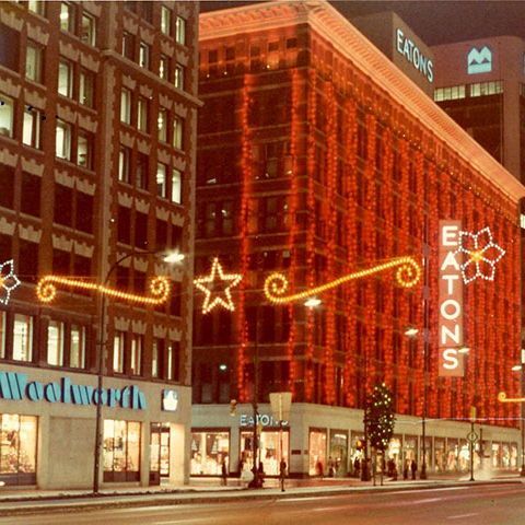 This vintage photo shows a nighttime city street, likely mid-20th century. A large brick building glows with red neon outlines and a prominent "EATONS" sign, indicating Eaton's department store. Festive lights form swirls, stars, and flowers across the facade and street, evoking holiday cheer.

To the left is a "Woolworth" store with blue signage and window displays. The wet street reflects lights, with few pedestrians and a Christmas tree visible. Background includes taller buildings with "EATONS" and "BMO" signs. The image has a warm, grainy nostalgic feel, watermarked "GRP" in the corner.