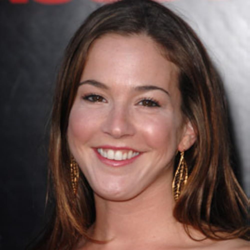 A photo of a woman with long, wavy brown hair, wearing a red strapless dress and dangling gold earrings. She is smiling and standing against a dark background with red and light-colored elements.