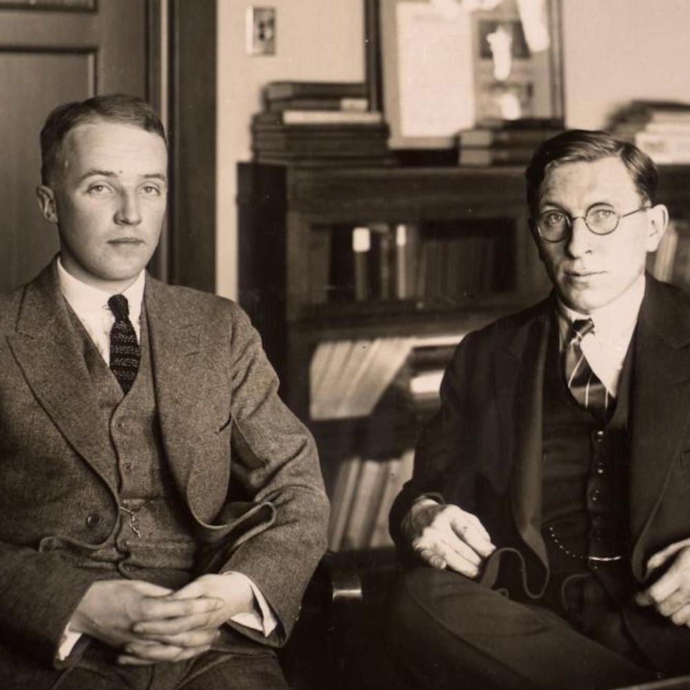 The black-and-white photograph shows two men, identified as Charles Best and Frederick Banting, seated side by side in a formal setting. Both are dressed in suits with ties, typical of early 20th-century professional attire. Charles Best, on the left, wears a three-piece suit with a patterned tie and has his hands clasped in front of him. Frederick Banting, on the right, is dressed in a darker suit with a striped tie and holds a pair of glasses in one hand while resting the other on his lap. Behind them is a wooden bookshelf filled with books and papers, along with a framed picture on the wall.
