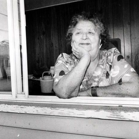 Black and white photo of Mary Two-Axe Earley leaning on a windowsill with her chin resting on her hand. She is wearing a floral-patterned dress with short sleeves. The window behind her shows a view of houses and a street outside. Inside, there is a wooden paneled wall, a chair, and a bucket on a table. The windowsill and exterior wall show signs of wear and peeling paint.
