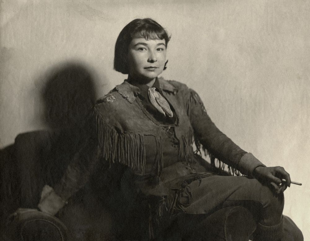 The black-and-white photo shows Anahareo sitting on a chair, dressed in a fringed leather outfit with a scarf at her neck. She holds a cigarette in her right hand, with a shadow cast on the wall behind her. The overall tone suggests a vintage, possibly early 20th-century setting.