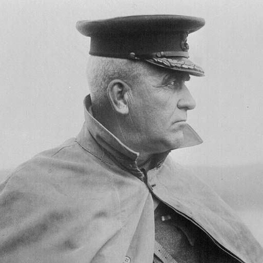 Black-and-white photograph showing a close-up side profile of an older man with short hair, facing right with a stern expression, wearing a dark peaked cap adorned with embroidery and an emblem, and a light-colored cape or overcoat draped over his shoulders, set against a blurred outdoor horizon.