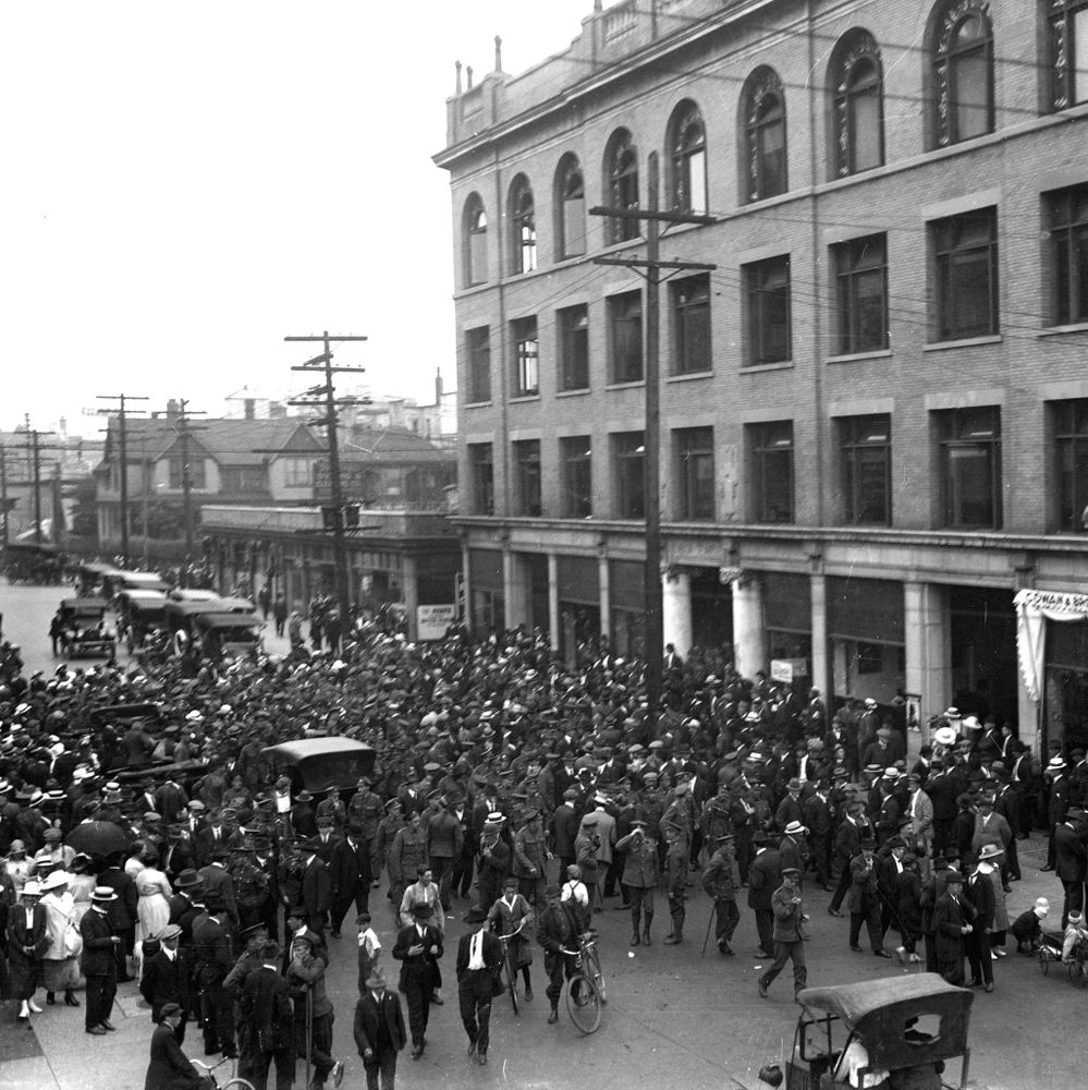 Black-and-white historical photograph of a large crowd of people, mostly men in hats and coats with some in military uniforms, gathered on a city street in front of a multi-story brick building with arched windows and signs reading "Cowan & Brookhouse" and others; vintage automobiles, utility poles, and streetcars are visible in the background.